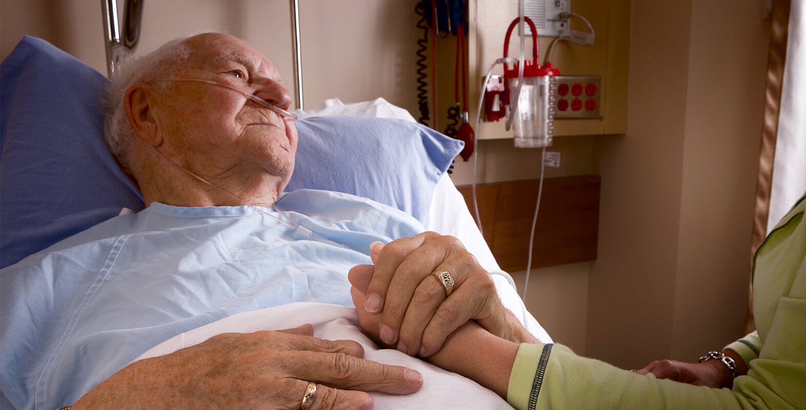 Elderly man in hospital bed holding someone else's hand representing palliative care.