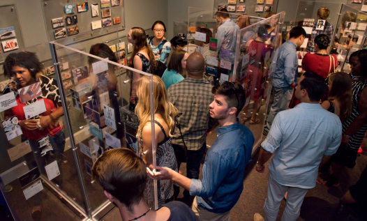 A group of people stand in a museum gallery filled with anonymous postcard confessions.