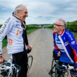 Two elderly cyclists in cycling gear standing on the road talking.