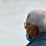 Elderly man with white hair facing away from camera and wearing a blue face mask pushed down.