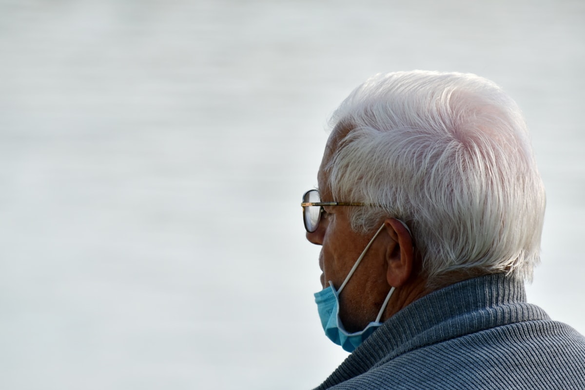 Elderly man with white hair facing away from camera and wearing a blue face mask pushed down.