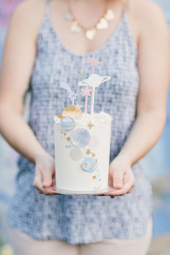 A woman holds a tall white cake decorated with stars and planets, the perfect offering for a celestrial remembrance event