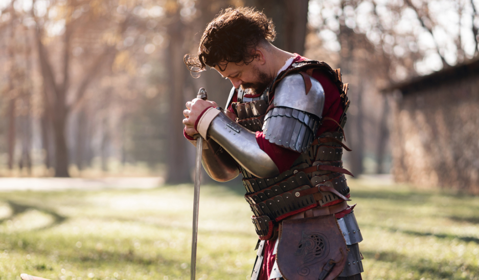 A man in a medieval knight costume is kneeling in a field while grasping the pommel of his sword and bowing his head in grief, mourning the death of a compatriot. His shield lies before him on the ground, and his helmet has been removed revealing curly brown hair.