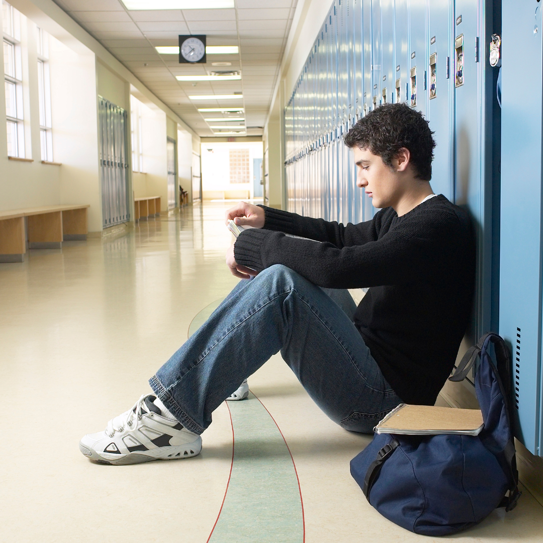 A teenage boy sits with his back against the lockers of a school hallway looking sad