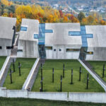 A series of concrete buildings studded with rectangular windows are situated in a row next to a field of black crosses. These buildings are the Mausoleum of the Martyrdom of Polish Villages, a monument to the innocent people lost in Nazi pacification campaigns.