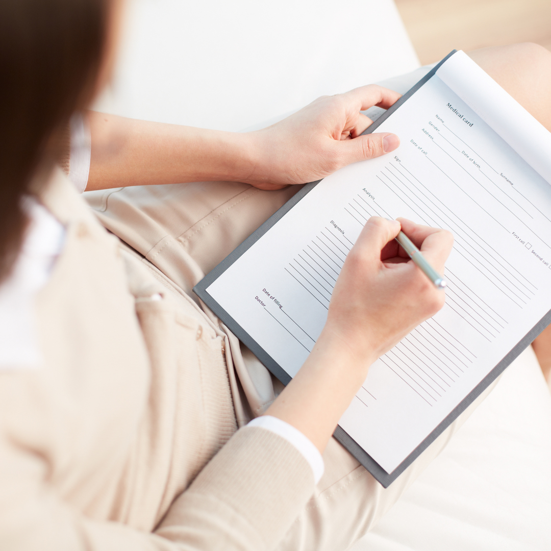The camera looks over the shoulder of a woman as she fills out medical paperwork while sitting in a hospital waiting room