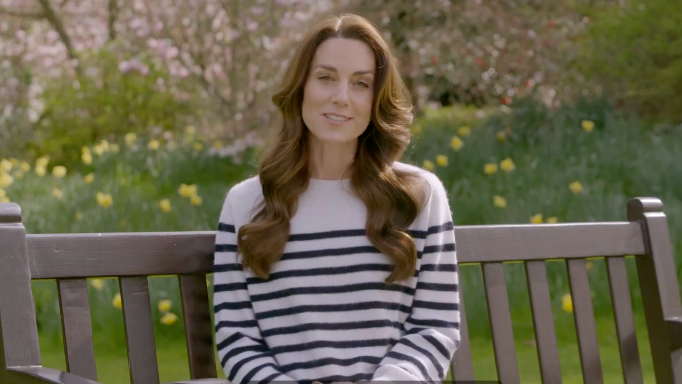 Kate Middleton, the Princess of Wales, sits on a sunny park bench wearing a cream-and-black stripped sweater as she issues a statement regarding her recent surprising cancer diagnosis
