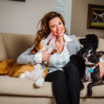 Dr. Dani McVety sits on a couch surrounded by three dogs. One is licking her chin while the other two gaze up at her.