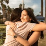 Two women embrace tearfully, with tropical trees in the background.