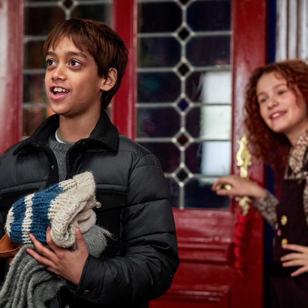 A smiling girl with curly hair in overalls closes her front door while her friend removes his winter hat and looks on in awe