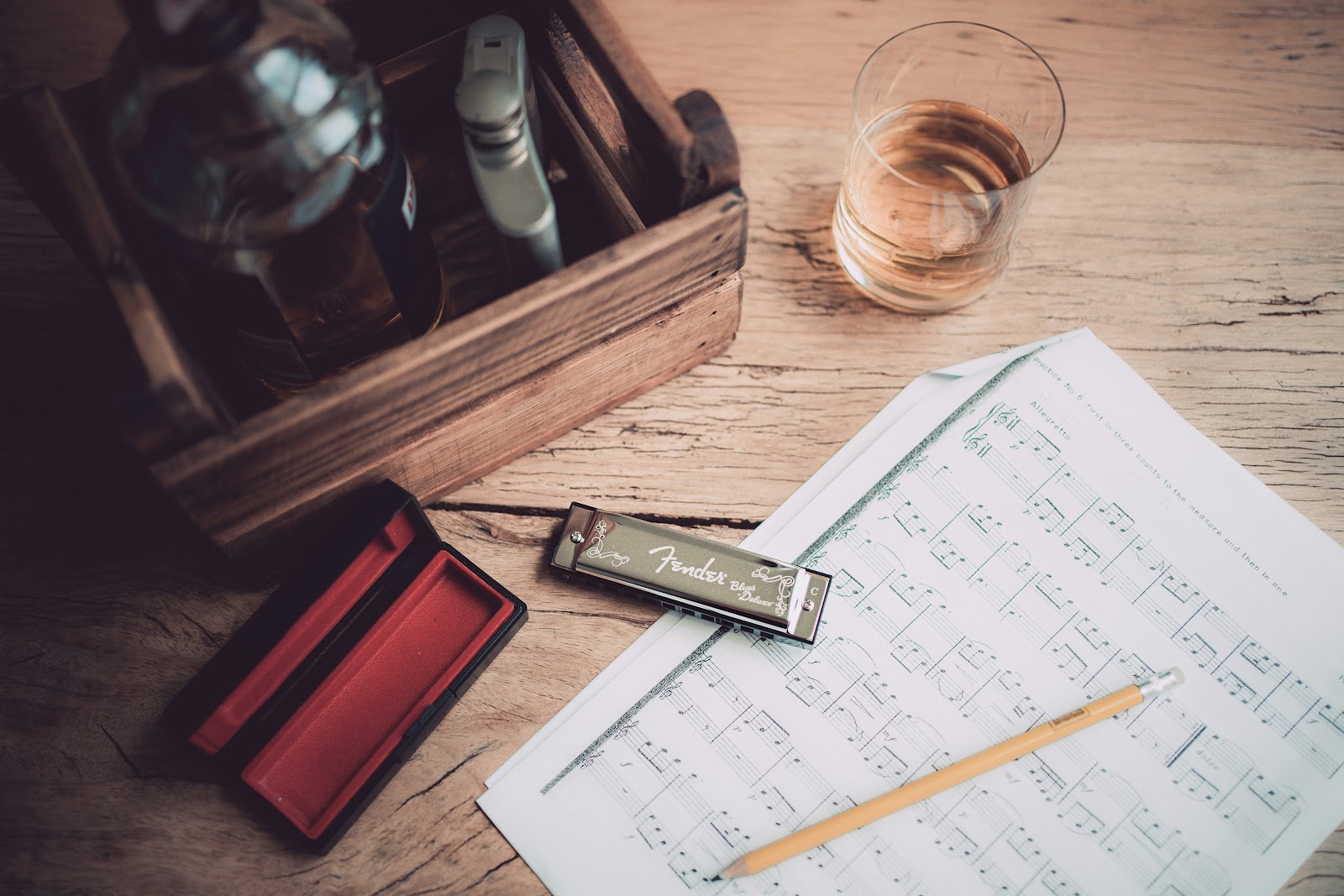 Several items are scattered across a wooden surface, including sheet music, a pencil, a harmonica, and an open harmonica case