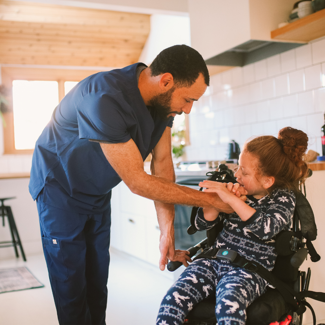 A male nurse in navy blue scrubs holds the hands of a young patient who is in a wheelchair