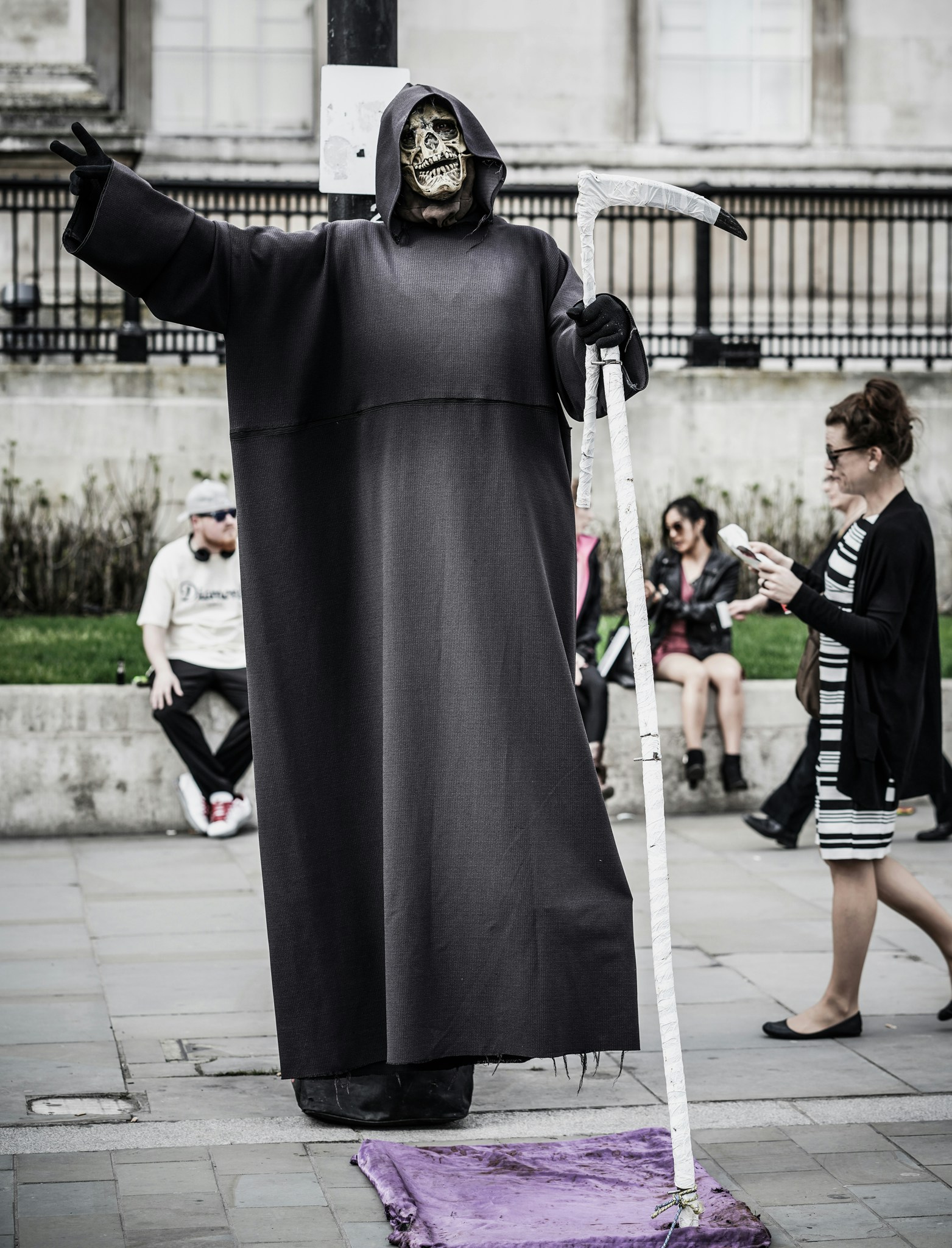 A person dressed as the Grim Reaper seems to be smiling and giving the "peace" sign with one hand while the other holds his scythe. People are walking past him on a city street, not noticing him at all.