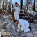 Two archaeologists in white protective clothing sort through debris on the foundation of a home destroyed by fire.