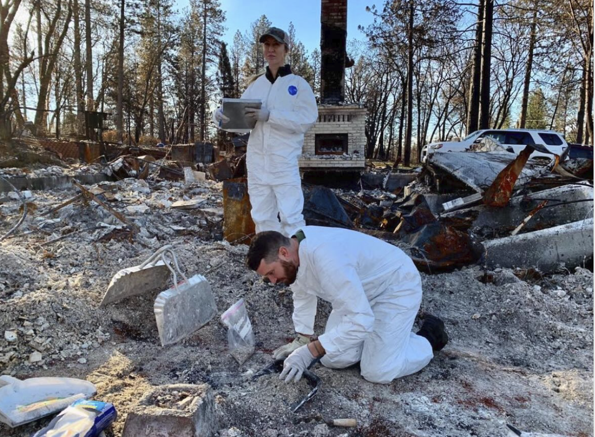 Two archaeologists in white protective clothing sort through debris on the foundation of a home destroyed by fire.