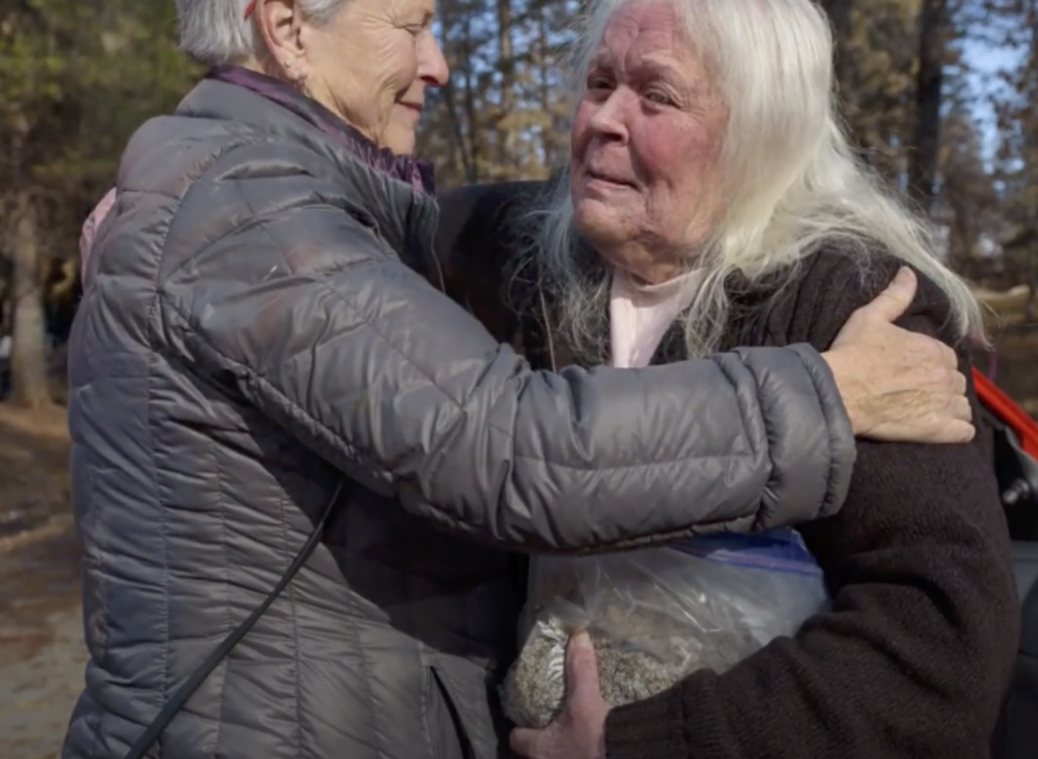 Two older women hug as one of them clutches the cremains of a loved one in a plastic bag. 