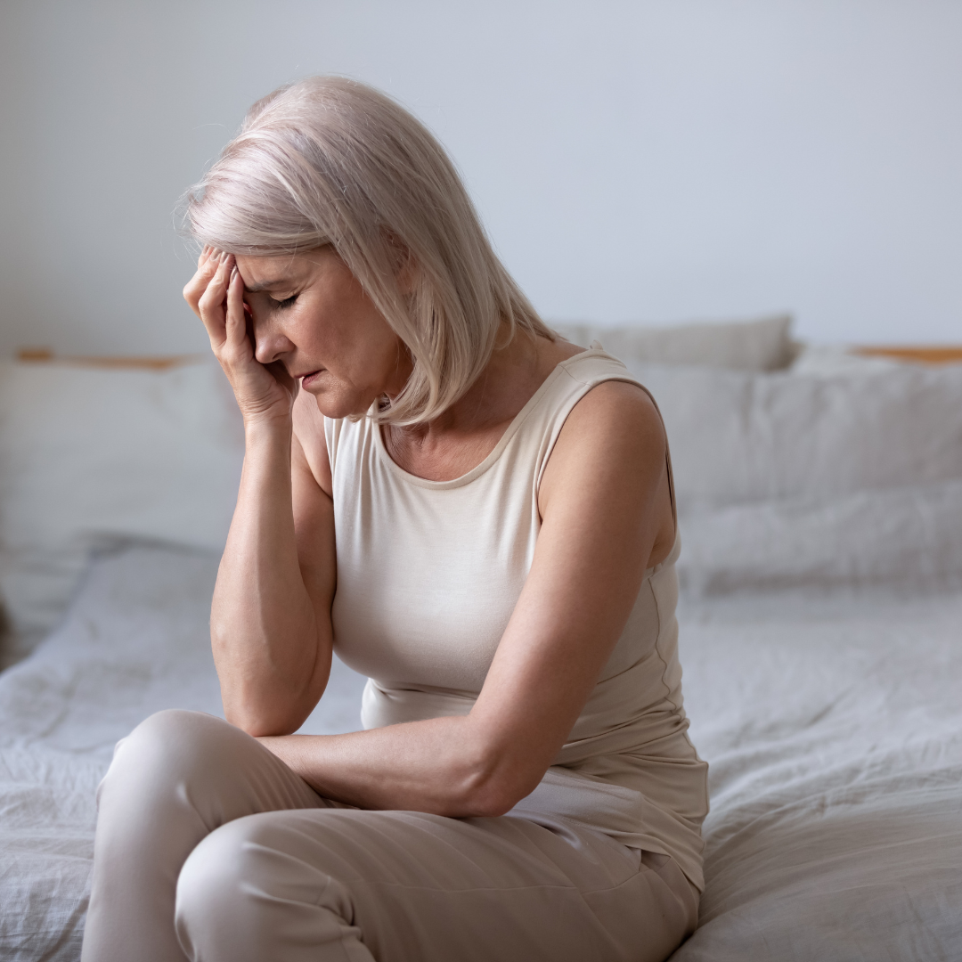 An older woman wearing all neutral tones sits on an equally neutral-toned bed, her head in her hands as if she's in pain.