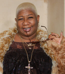 A black woman with short blonde hair smiles winningly while biting down on her fashion glasses