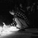 A man in a plaid shirt crouches down to light a candle at a vigil