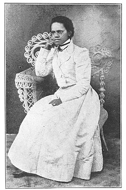 A grainy, black and white photograph of a young, Black woman sitting in an ornate chair with her chin propped on her fist. She is dressed in a white dress, in fashion common to the early 1900s.
