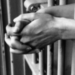 A black-and-white photograph of an inmates' hands clasped through the bars of their cell.