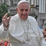 Pope Francis, a kind-faced older gentleman, waves from an elevated position to the crowds clamoring for attention. He is wearing white Catholic Pope vestments, and a silver cross on a chain around his neck.