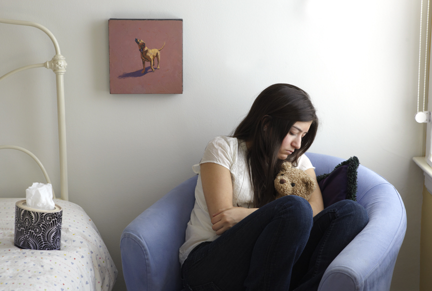 a woman sitting in a blue chair in a hospital room holding a teddy bear grappling with death and mortality