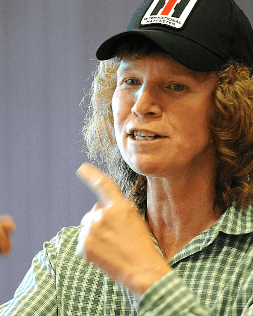 A close-up photograph of Deborah Lipsky, a woman with short, voluminous hair tucked under a black baseball cap, is speaking, gesturing with her hands.