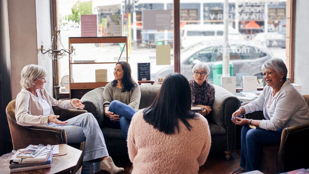 A group of women of varying ages are sitting in comfortable couches and chairs at a cafe, discussing death in a frank manner.