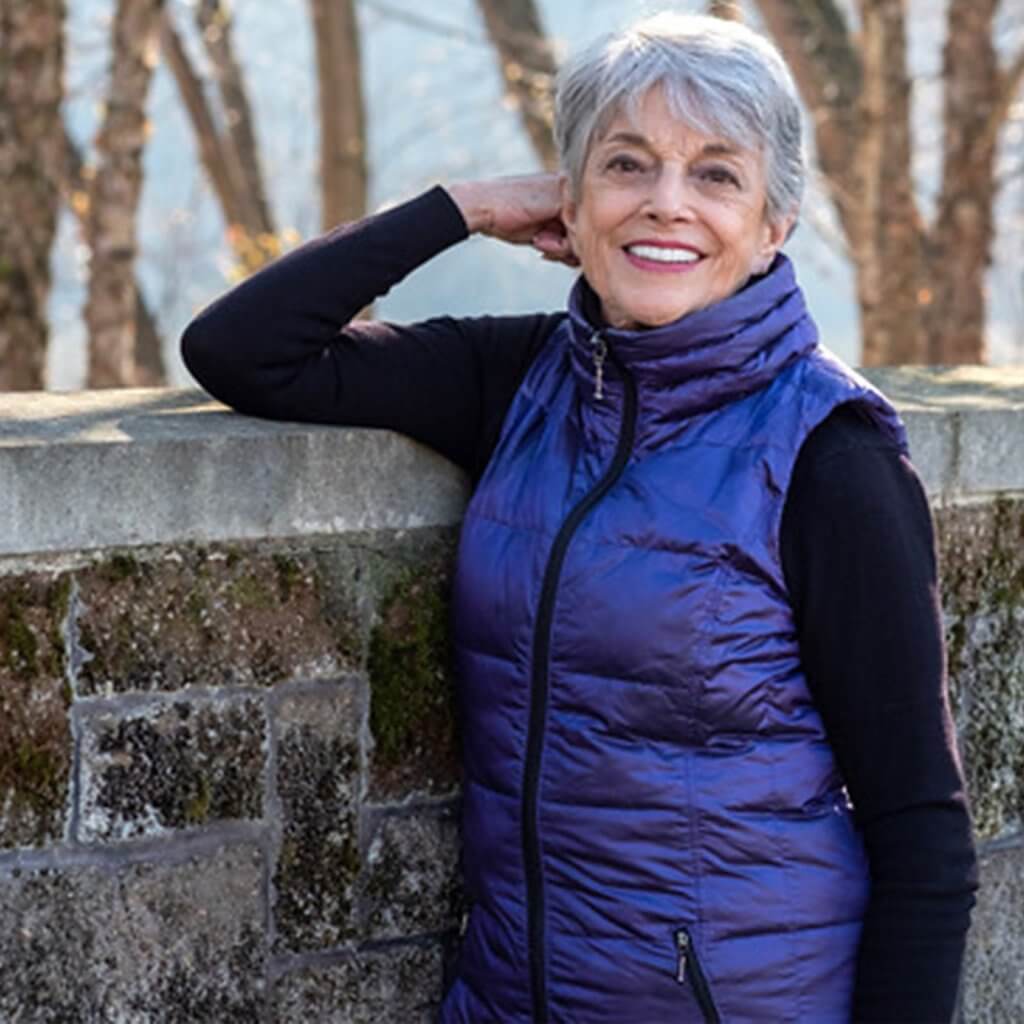 A photograph of the author of "The Art of Grieving," Sheila K. Collins. She is standing against a stone wall with trees in the background.