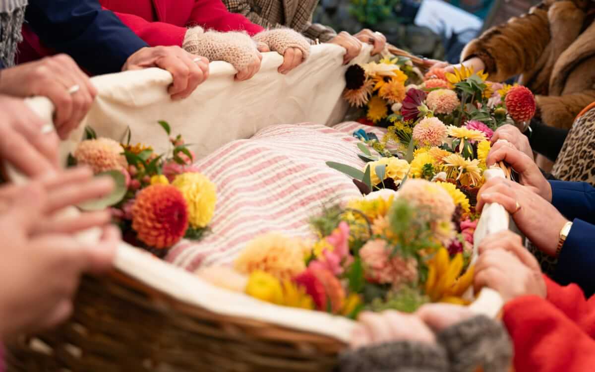 Several hands are shown holding a casket filled with flowers and blankets.