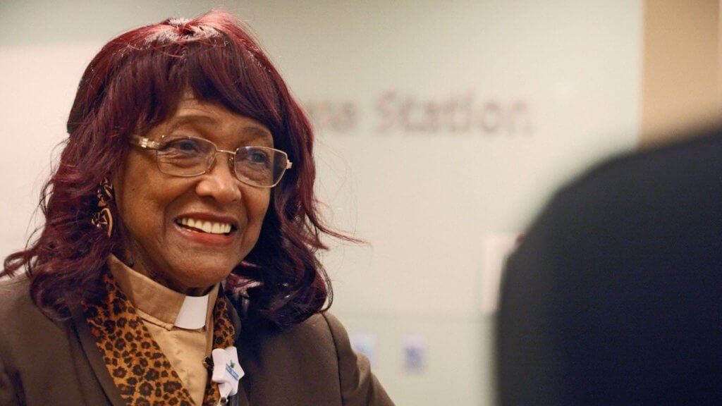 An older African American woman wearing a clerical collar and gold-rimmed glasses smiles as she's talking with someone just out of frame.