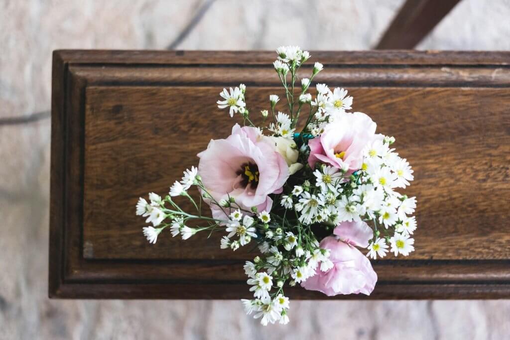 A closeup image of a closed brown casket with a light pink floral arrangement on top of the lid.