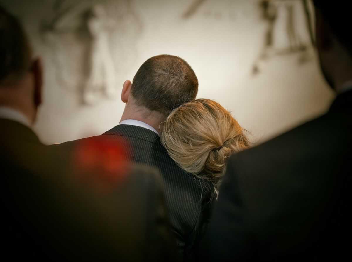 Two people leaning their heads against each other, facing away from the camera. They are wearing formal attire, and it seems they are attending a funeral or memorial service.