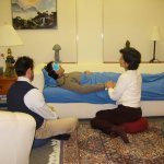 In a spa-like room, a patient is lying on a low couch/bed, wearing a light blue eye mask and a light blanket is drawn up to their chest. Holding the patient's hand is a woman sitting on a cushion, and a third person sits on a cushion nearby.