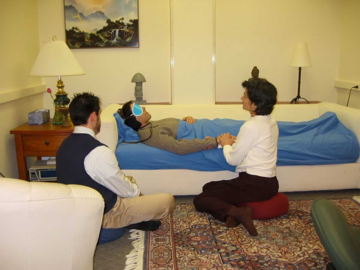 In a spa-like room, a patient is lying on a low couch/bed, wearing a light blue eye mask and a light blanket is drawn up to their chest. Holding the patient's hand is a woman sitting on a cushion, and a third person sits on a cushion nearby.