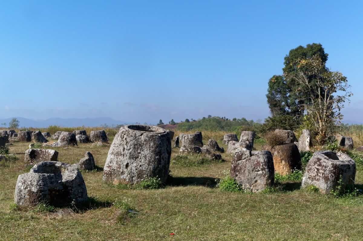 Dozens of stone megaliths shaped like cups or jars dot a broad field with few trees and a few shrubs; these artifacts are mortuary urns on Laos's Plain of Jars.