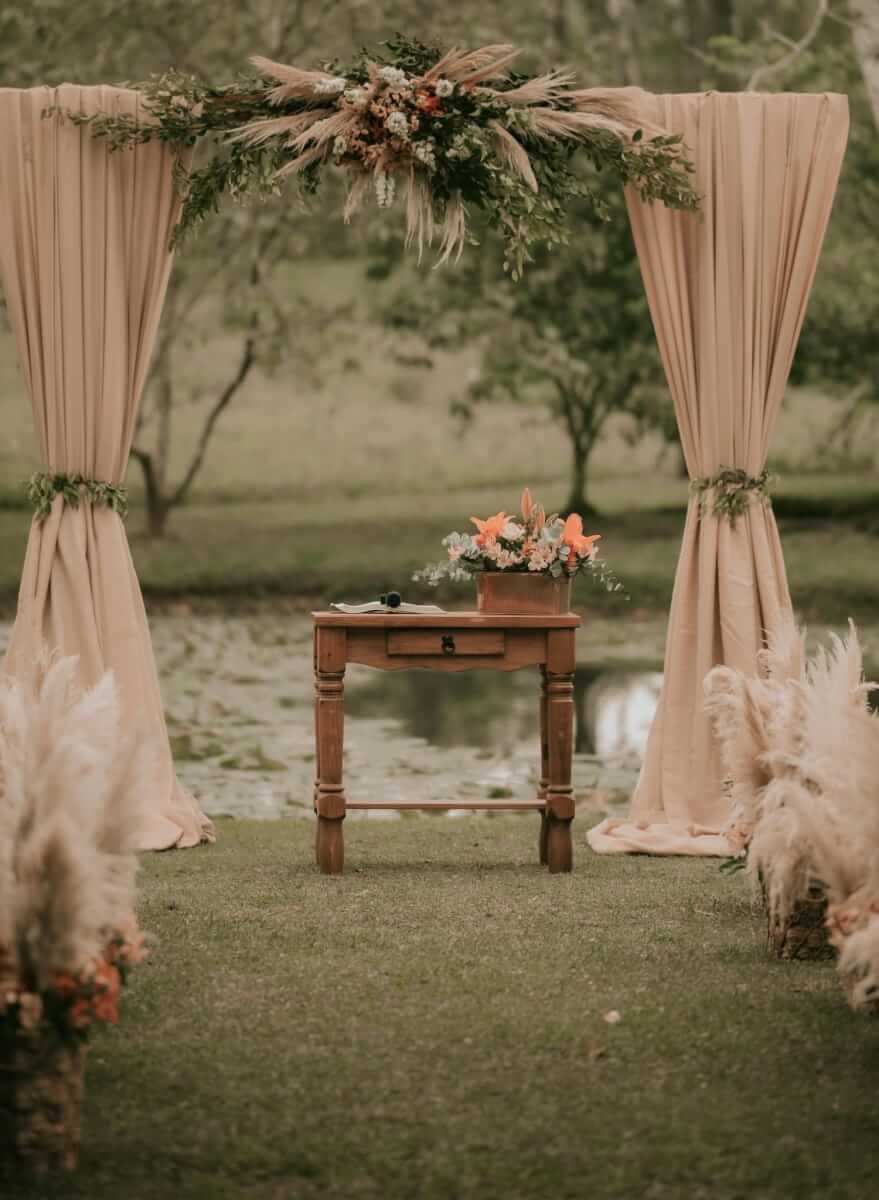 A wooden arbor is beautifully decorated at the end of a casual aisle in a field with a pond as a background. There is soft, rose-colored fabric draped over the arbor, and floral arrangements with fall blooms and pampas grass serve as the centerpiece, as well as decorations along a casual aisle, as a setting for a memorial service or eulogy.