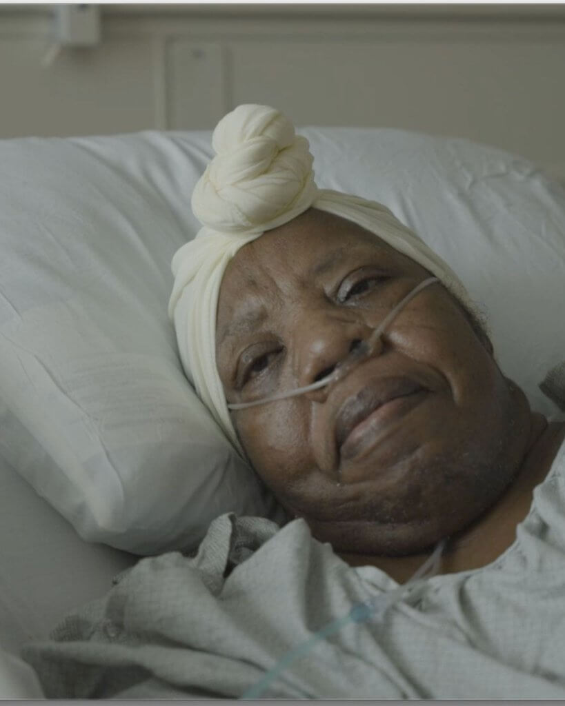 An older African American woman wearing a knotted headscarf and an oxygen cannula in her nose is looking at someone offscreen from her hospital bed. 