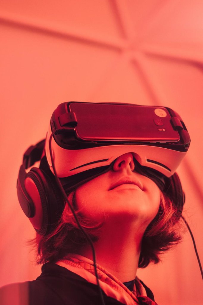 A close-up, low-angle shot of a person looking upward while wearing a white smartphone-powered virtual reality headset and large black over-ear headphones. The entire scene is bathed in a vibrant, monochromatic red light.