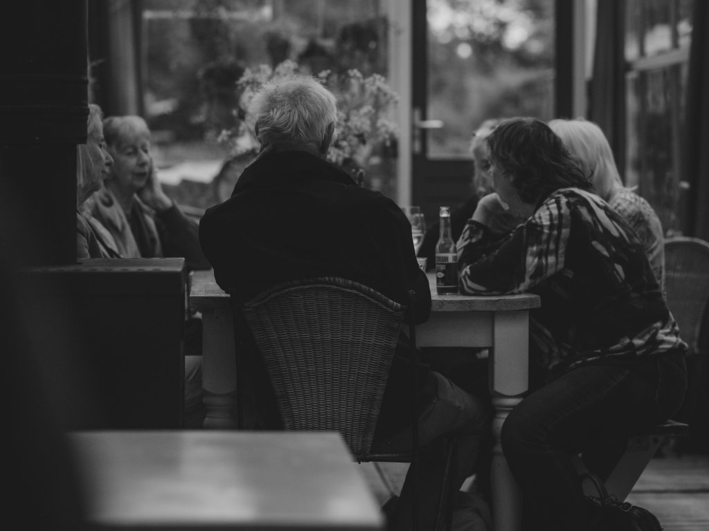 A black and white photograph shows a group of older adults gathered around an outdoor or sunroom table, seen from a low angle with a blurry foreground object. A man with white hair sits with his back to the camera in a wicker chair, while several women sit across and beside him engaged in conversation. A glass bottle and drinks sit on the wooden table.