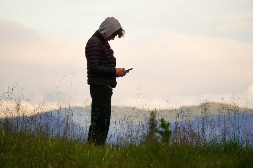 A side profile of a person standing isolated in a desolate, grassy field, huddled in a dark puffer jacket with their hood up as they look down at a smartphone. The background of cold, hazy mountains and a muted sky creates a somber mood of disconnection and anxiety.