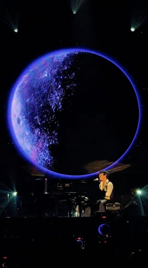 Bruno Mars performs at a piano beneath a large glowing blue moon during a live concert performance of “Talking to the Moon.”