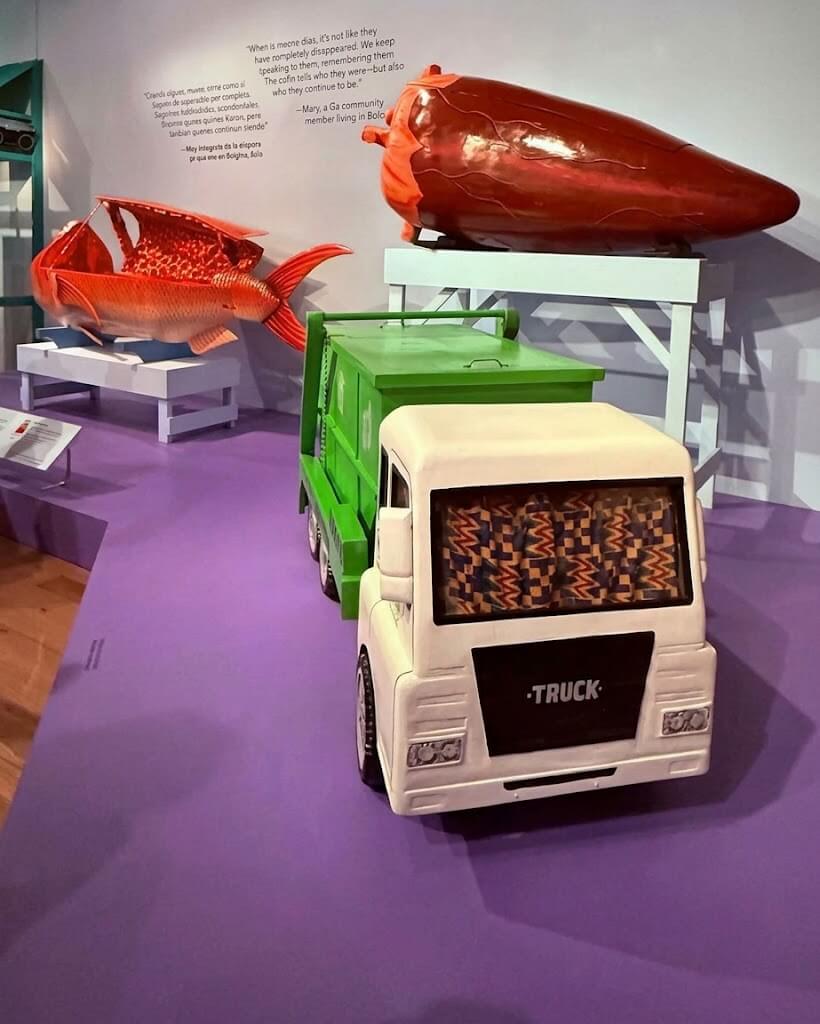 A museum exhibition displaying three Ghanaian fantasy coffins shaped like a white truck, a bright red fish, and a red chili pepper.