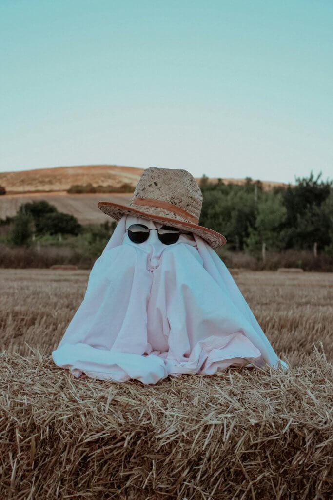 Ghost costume wearing sunglasses and a straw hat on a hay bale in a field for a Halloween-themed celebration of life
