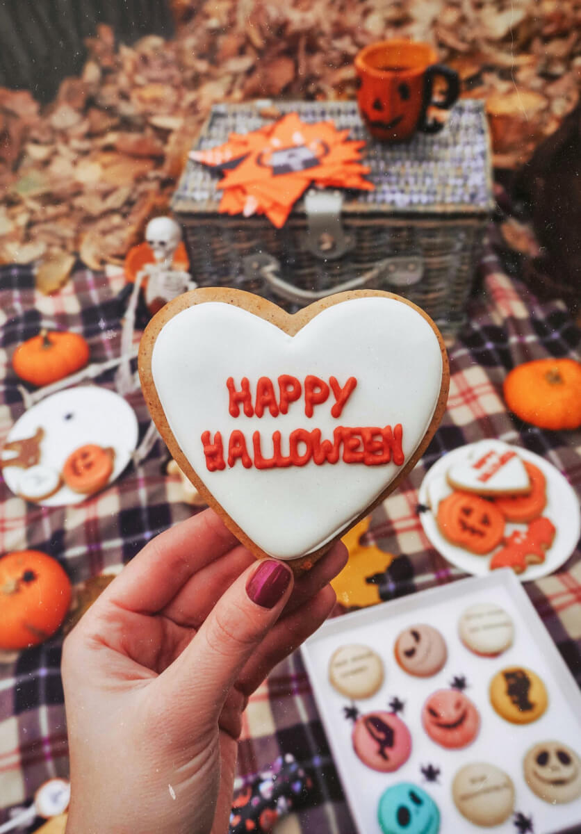 Hand holding a heart-shaped Halloween cookie surrounded by decorated Halloween cookies and fall decorations