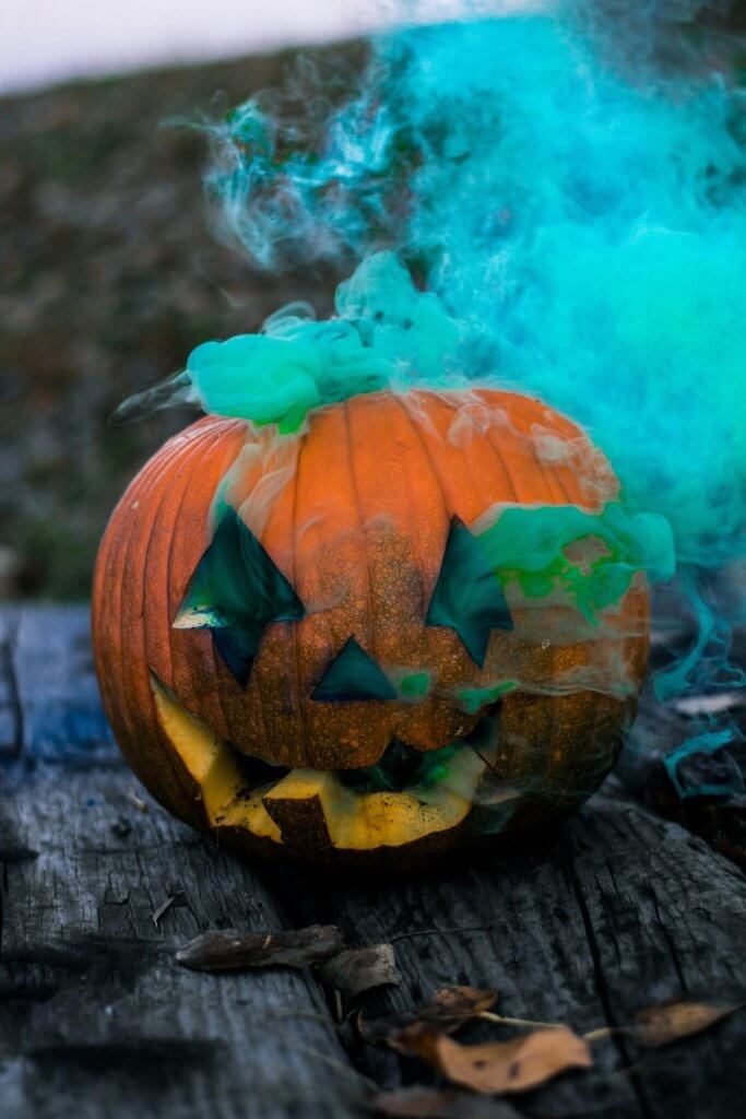 Carved jack-o’-lantern surrounded by blue and green smoke on a wooden table for a Halloween-themed celebration of life