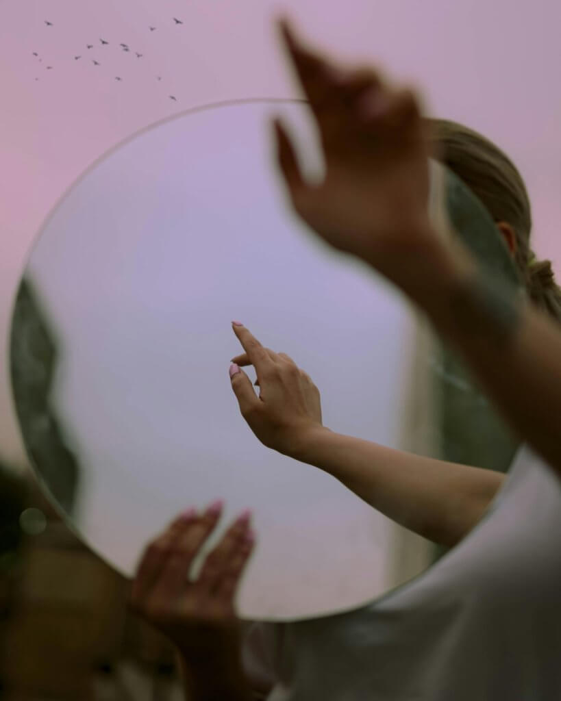 Person holding a round mirror against a soft pink sky, one hand reaching upward as a flock of birds flies overhead, evoking longing and letting go