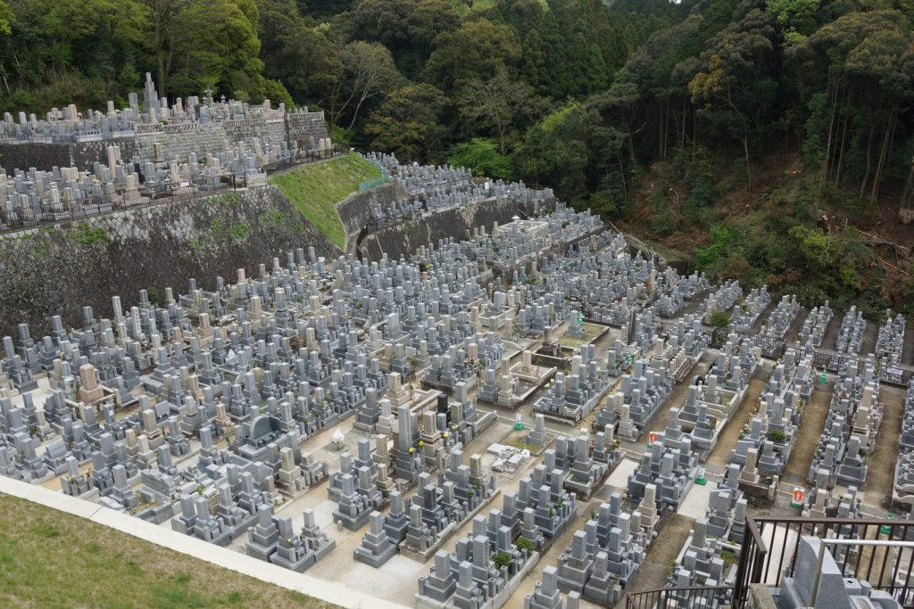 Aerial view of a Japanese hillside cemetery with terraced rows of granite grave markers and a forested slope behind