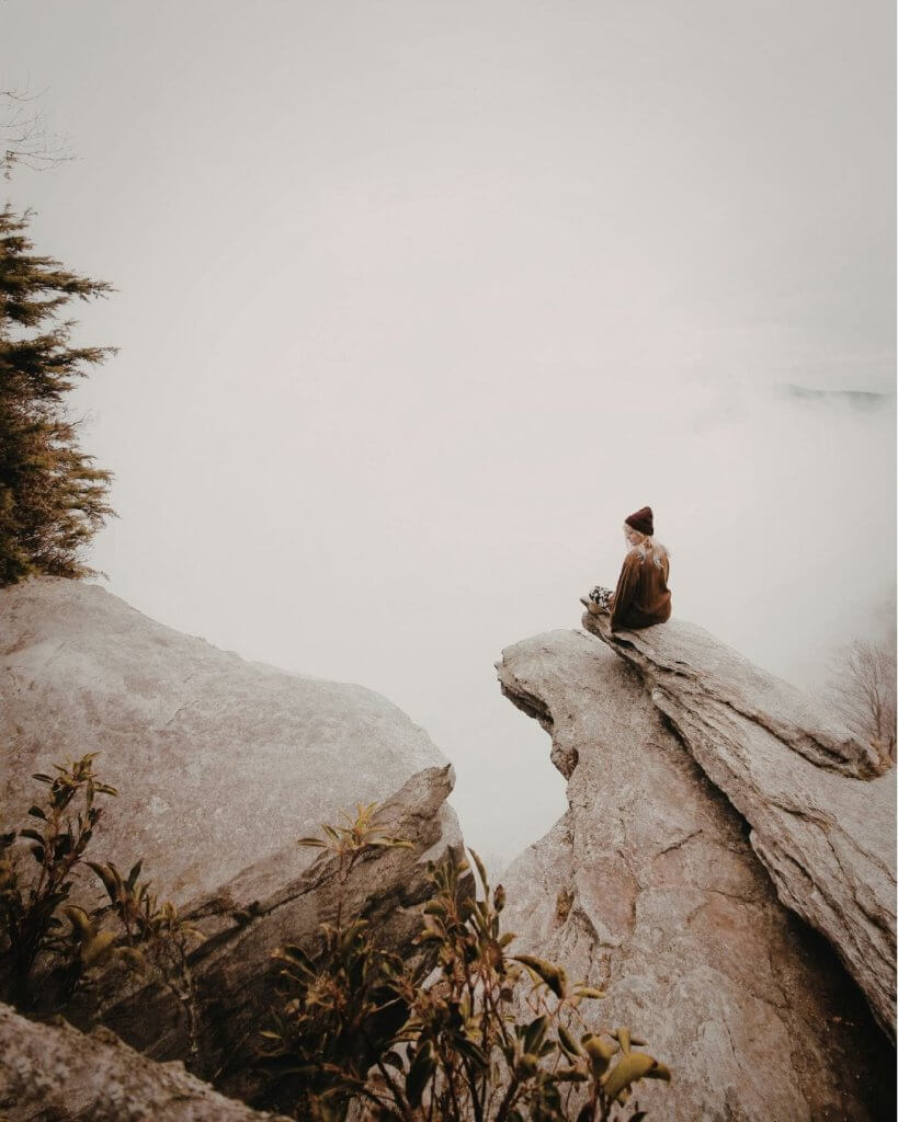 A person sits alone on a rocky mountain outcrop overlooking a fog-covered landscape, evoking solitude and reflection in nature.